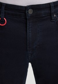 Ciemnoniebieskie dżinsy z guzikiem, z szlufkami na pasek oraz czerwoną tkaniną jako akcentem na przednim pasku.