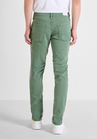 Jeans verdes de corte recto, fabricados en algodón, con bolsillos traseros, textura suave, diseño minimalista, combinados con zapatillas blancas, mostrados desde la parte posterior.