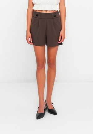 Shorts marron ajustés avec des plis sur le devant et deux boutons noirs à la taille. Tissu lisse avec une coupe structurée, associés à des talons noirs.