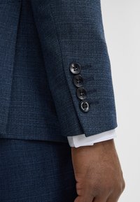 Manche de costume bleu marine texturée avec quatre boutons noirs, poignet de chemise blanche et main d'une personne à la peau foncée au repos.