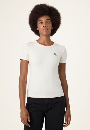 Donna con acconciatura afro naturale che indossa una maglietta aderente a maniche corte color avorio con un piccolo logo sul petto e pantaloni neri.