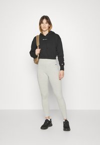 Donna che indossa un hoodie nero corto, leggings grigio chiaro, sneakers nere e porta uno zaino beige su una spalla, in piedi su uno sfondo bianco.