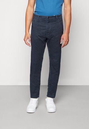 Chinos in cotone blu navy con vestibilità slim, dotati di chiusura anteriore con bottone, passanti per cintura e tasche laterali, abbinati a sneaker bianche.