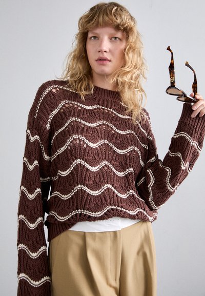 Brun strikket sweater med hvide bølgemønstre, rund halsudskæring og lange ærmer. Modellen holder solbriller i skildpaddemønster.