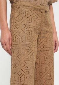 Pantalon marron à motifs avec des découpes géométriques et un tissu texturé, avec des coutures visibles et une taille haute.