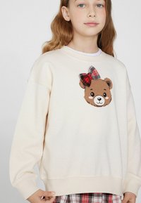Sweatshirt crema con grafica di un volto di orso bruno, decorata con un fiocco a scacchi rosso e nero, con polsini e orlo a coste.