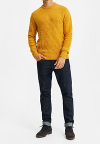 Stickad gul tröja med ribbstickade och flätade texturer, rund halsringning, kombinerad med mörka denimjeans och mörka skor.