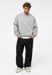 Grijze oversized sweatshirt met geribbelde manchetten, gecombineerd met zwarte, loszittende jeans. Lichtgekleurde sneakers met mesh- en rubberaccenten.