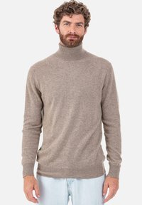 Pull-over à col roulé marron en matière douce avec une coupe ajustée, poignets et bordures côtelés. Porté avec un jean bleu clair.