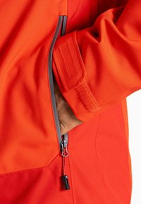 Rote Outdoor-Jacke mit einer seitlichen Reißverschlusstasche, ausgestattet mit einem strukturierten Stoff und schlanken Linien, ergänzt durch einen kontrastierenden Reißverschluss.