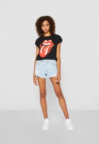 Sort t-shirt med et stort rødt Rolling Stones-logo, parret med slidte lyseblå denim shorts og hvide sneakers.