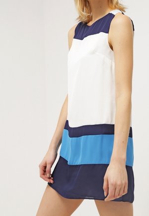 Mouwloze colorblock-jurk met marineblauwe, witte en felblauwe horizontale panelen gedragen door een staande vrouw tegen een effen achtergrond.