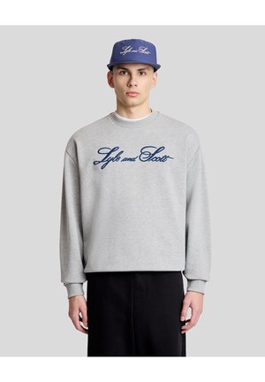 Ung mand iført en grå sweatshirt og en blå kasket, begge med "Lyle and Scott" broderet i blåt skrift, stående foran en grå baggrund.