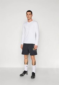 Jovem vestindo uma camisa branca de manga comprida, calções pretos da Puma, meias brancas da Puma e sapatilhas pretas, em pé contra um fundo simples.