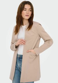 Veste beige cintrée avec un design structuré, manches longues et poches avant, portée sur un haut blanc et un jean bleu. Texture de tissu lisse.