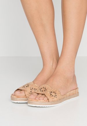 Pieds portant des sandales à enfiler en liège beige avec un motif floral découpé et une semelle blanche texturée sur un fond de sol clair.