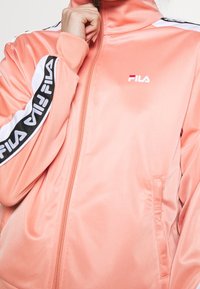 Peach-colored Fila zip-up jacket with black and white stripes on sleeves. Smooth texture, high collar, and logo on the chest.