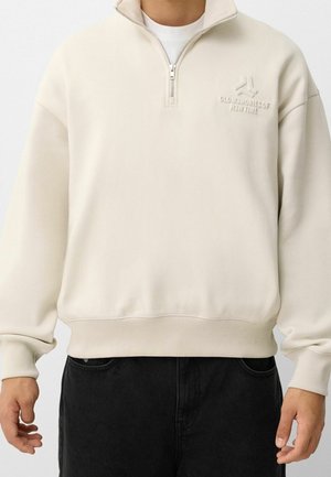 Man wearing off-white half-zip sweatshirt with embossed text "Old Memories of Best Time" and black pants, hands relaxed by sides.