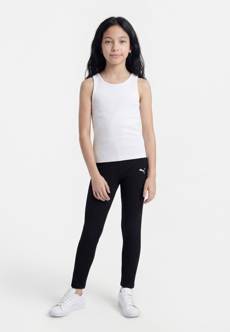 Fată stând în picioare cu păr lung și negru, purtând un top alb fără mâneci, colanți negri Puma și adidași albi pe un fundal alb simplu.