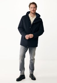 Veste à capuche marine avec boutons sur le devant, poches latérales et finition lisse. Portée sur une chemise de couleur claire, associée à un jean gris et des bottes noires.