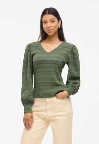 Pull en tricot vert avec un décolleté en V, motif texturé et manches bouffantes. Poignets ajustés, porté avec un pantalon de couleur claire.