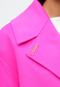 Lys pink uldfrakke med en bred krave og en gul "Future" brosche accent på klappen, der har en blød tekstur.