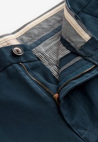 Donkergroene broek met ritssluiting en knoopsluiting. Binnenkant heeft een gestreepte en patroonvoering. Gladde stof met een getailleerde pasvorm.