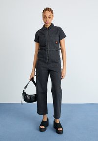 Mørk denim jumpsuit med korte ermer, glidelås foran og brystlomme. Brukt med plattform sko og en liten svart håndveske.
