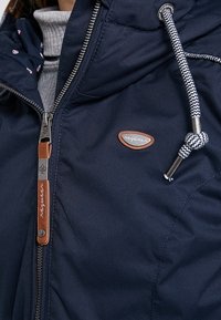 Navy blue insulated jacket with a zip front, drawstring hood, and leather accents. Features patterned lining and logo patch on the chest.