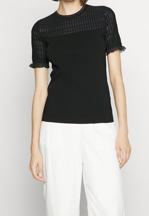 Kobieta w czarnej koszulce z krótkim rękawem, przezroczystymi paskowanymi rękawami i falbankami, w połączeniu z białymi spodniami na jednolitym tle.