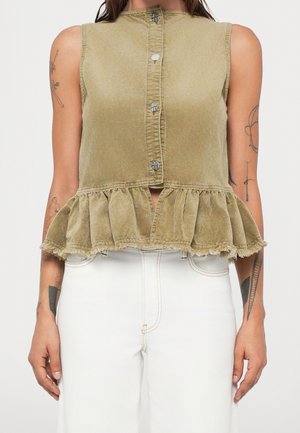 Haut sans manches en velours côtelé vert olive avec boutons sur le devant et ourlet à volants, porté avec un pantalon blanc taille haute, montrant des tatouages sur les bras.