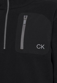 Svart fleecejacka med grå vertikal dragkedja på bröstet och vit "CK"-logotyp broderad under fickan.