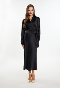 Abito avvolgente in satin nero con maniche lunghe, vita cintata e gonna fluida. Abbinato a tacchi a punta metallici.