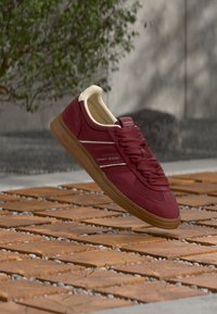 Bordeauxfarbener Sneaker mit glattem Wildleder-Obermaterial, cremefarbenen Akzenten, flachen Schnürsenkeln und einer strukturierten Kautschuksohle. "TOMMY JEANS"-Logo an der Seite.