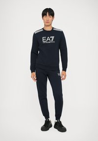 Completo di felpa e pantaloni in blu navy con dettagli del logo in bianco. La felpa presenta accenti a righe sulle spalle; i pantaloni hanno polsini elasticizzati.