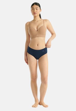 Vrouw staat blootsvoets in beige beha en marineblauwe onderbroek, houdt de tailleband vast, met neutrale expressie tegen een effen lichte achtergrond.