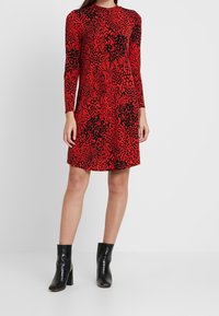 Robe longue à manches longues rouge avec un imprimé animalier noir audacieux, dotée d'un col rond et d'une silhouette décontractée. Portée avec des bottines noires.