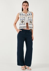 Mouwloos, wit geborduurd vest met blauwe en gele ontwerpen, gecombineerd met losse, donkerblauwe broeken en witte sandalen.