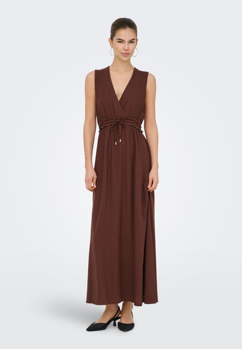 Femme portant une robe maxi marron sans manches avec un profond décolleté en V et une taille resserrée par un cordon, associée à des chaussures noires à talons bas et des boucles d'oreilles créoles.