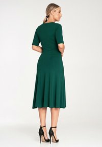 Zielona sukienka w odcieniu emerald z krótkimi rękawami, dopasowanym gorsetem, rozkloszowanym dołem i długością do kolan. W komplecie z czarnymi sandałami na kostkę.