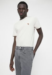 T-shirt di cotone beige chiaro con un piccolo logo animale ricamato, abbinato a jeans in denim grigi con chiusura a bottoni e tasche laterali.