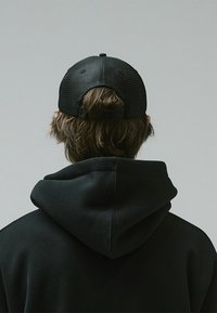 Black cap with textured fabric, adjustable strap, worn backwards, paired with a black hoodie featuring a visible hood and seam details.