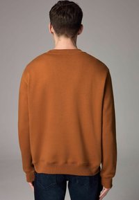 Sudadera marrón con cuello redondo y puños acanalados, que presenta una textura suave y un corte relajado. Combinada con jeans oscuros.