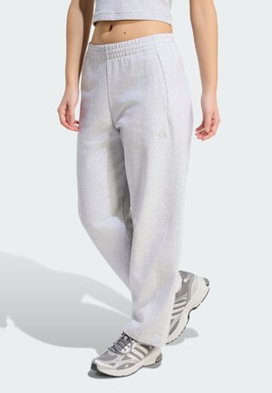 Osoba nosící světle šedé tepláky Adidas a šedé sportovní tenisky, stojící s jednou nohou mírně vpřed na světlém pozadí.