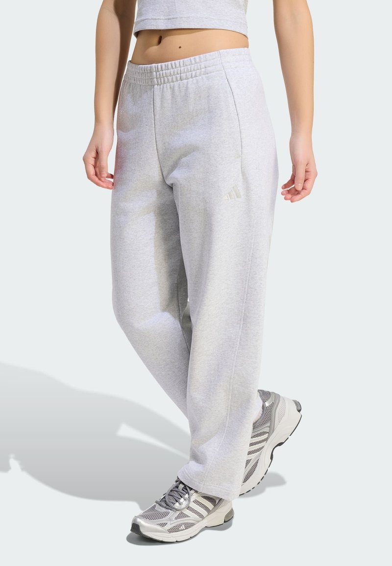 Personne portant un pantalon de jogging Adidas gris clair et des baskets de sport grises, debout avec une jambe légèrement avancée sur un fond clair.