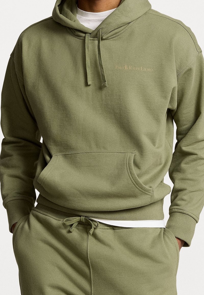 Homme portant un sweat à capuche vert olive et un pantalon de survêtement assorti, les mains dans les poches, t-shirt blanc visible en dessous, logo texte sur la poitrine.