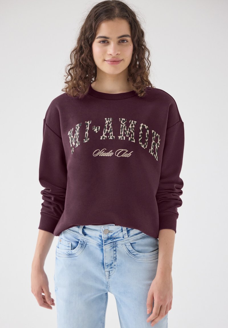 Jeune femme aux cheveux bouclés portant un sweat-shirt bordeaux avec le texte "MI'AMOR Studio Club" et un jean bleu clair sur un fond uni.
