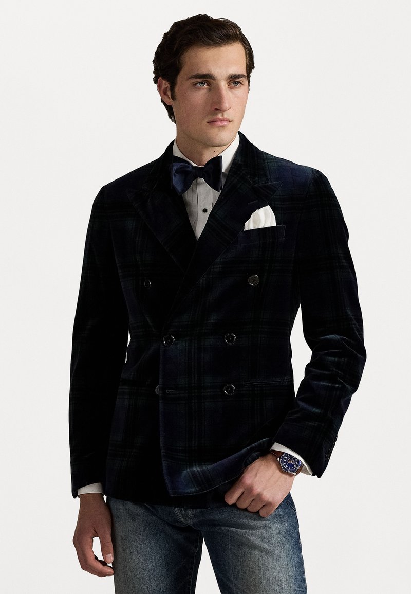Blazer en velours à double boutonnage en tartan bleu marine et vert, associée à une chemise habillée blanche, un nœud papillon noir et un jean ajusté.