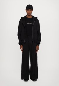 Svart zip-up jacka, svart grafisk t-shirt och vida svarta byxor. Slät textur, avslappnad passform och matchande svart keps med logotyp.