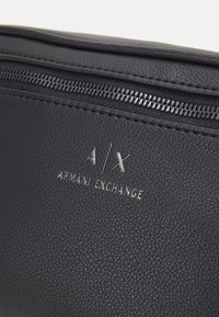 Schwarze, strukturierte Leder Tasche mit einem silbernen Reißverschluss und dem eingeprägten Logo "A/X ARMANI EXCHANGE" auf der Vorderseite.